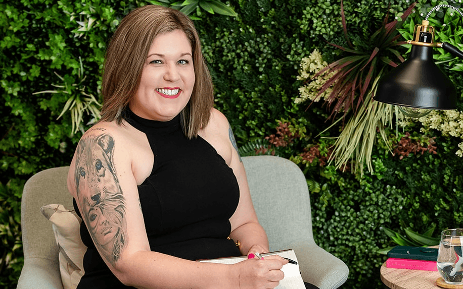 A woman sitting in a modern chair with a green plant wall behind her, smiling while writing in a notebook. She is wearing a black outfit and high heels.